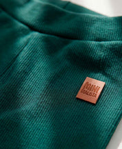 Calça Slim Canelada Bebê Liso Deep Green MiniMalista — cós elástico wide sem cadarço, etiqueta de couro caramelo no hip, malha canelada verde escuro 100% algodão fitted.
